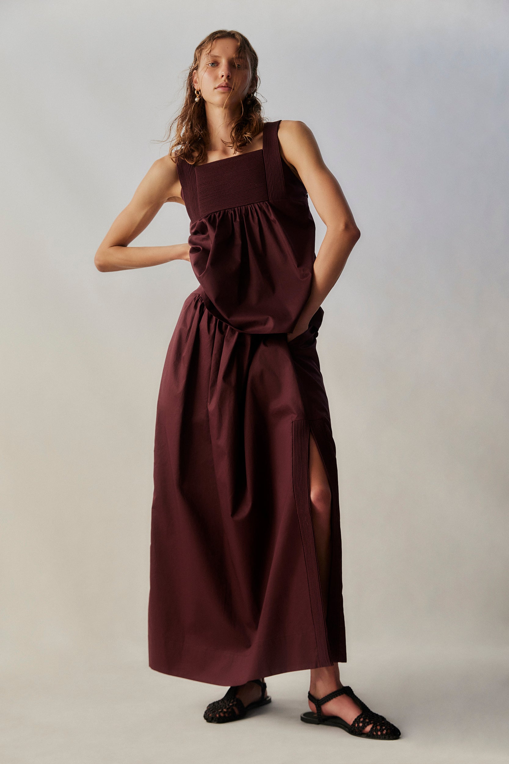 Woman wearing a long burgundy top and skirt with a slit on a plain background